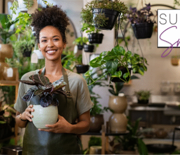 Small business owner smiling whilst holding her product, a plant. With the Support Small UK Logo in the top right hand corner.